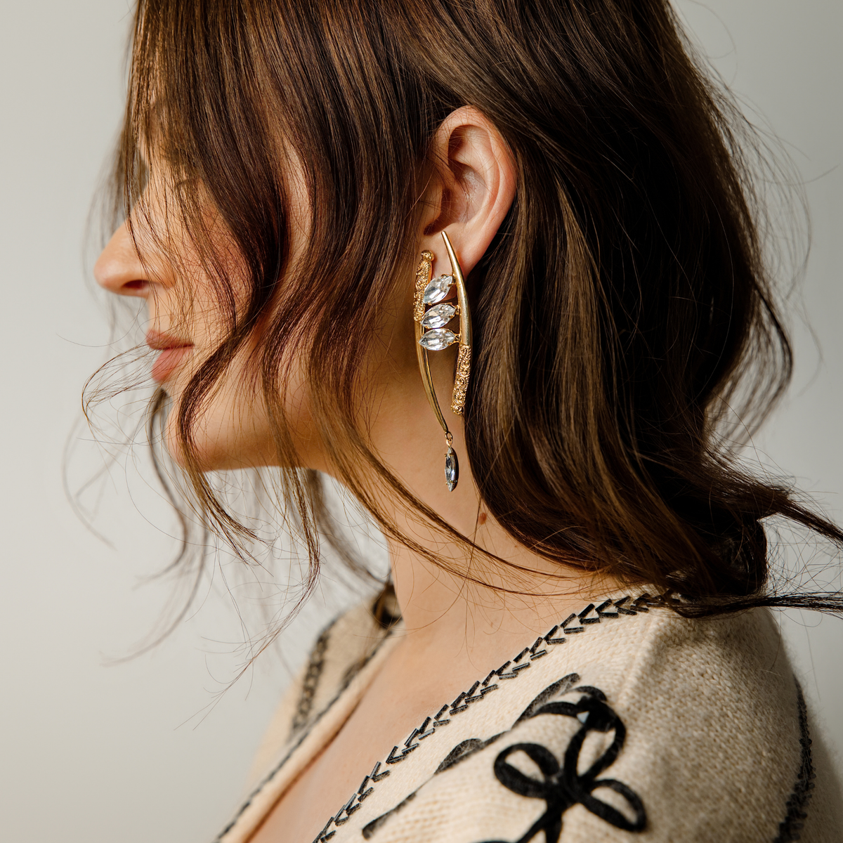 A close-up of a woman wearing a pair of asymmetric gold crescent-shaped drop earrings featuring marquise-cut clear, Swarovski crystals and ornate gold filigree details, set against a gray background with a cream knit top featuring black embroidery.