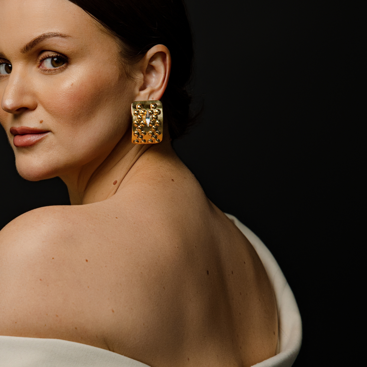 A close-up lifestyle shot of a woman wearing a chunky, gold square stud earring decorated with a raised cluster of small gold beads and a clear marquise-cut Swarovski crystal in the center against a solid black background.