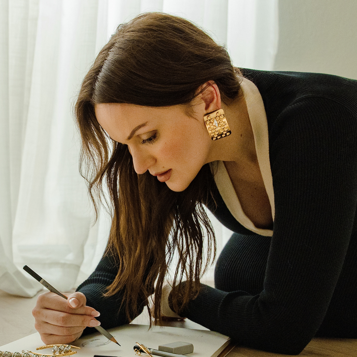 A woman sketching jewelry designs wearing a pair of large gold square stud earrings decorated with a raised cluster of small gold beads and a clear marquise-cut Swarovski crystal in the center.
