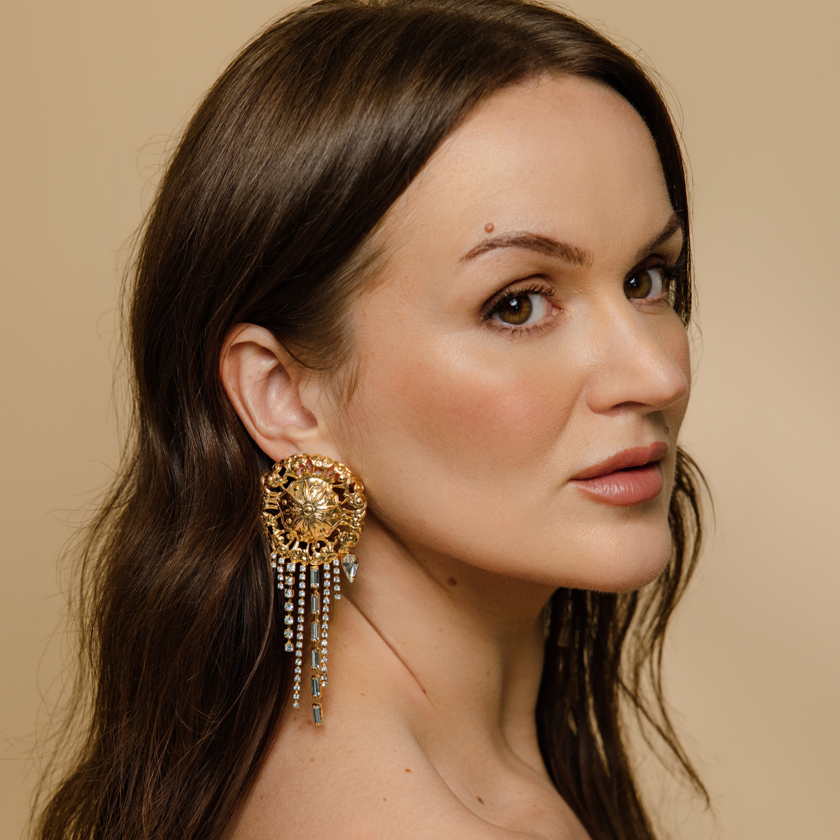 A close-up portrait of a woman wearing a large ornate gold filgree statement earring with dangling Swarovski crystals against a warm beige background.