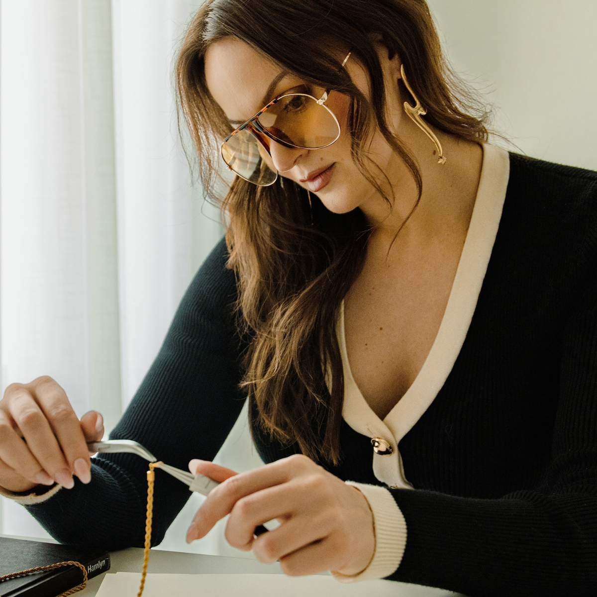 A woman with gold-rimmed glasses is shown in a close-up lifestyle shot, wearing a black ribbed sweater with cream-colored trim. She is making jewelry wearing gold sculptural earrings with a Swarovski crystal at the bottom.