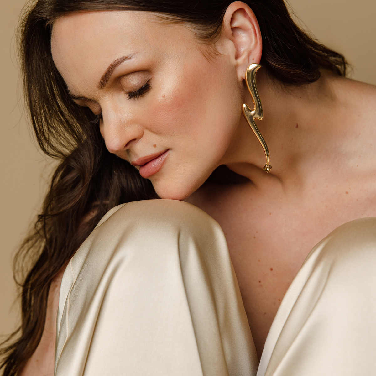 A close-up lifestyle shot of a woman wearing a large structural earring with a tonal Swarovski crystal at the bottom and a cream satin top against a warm beige background.