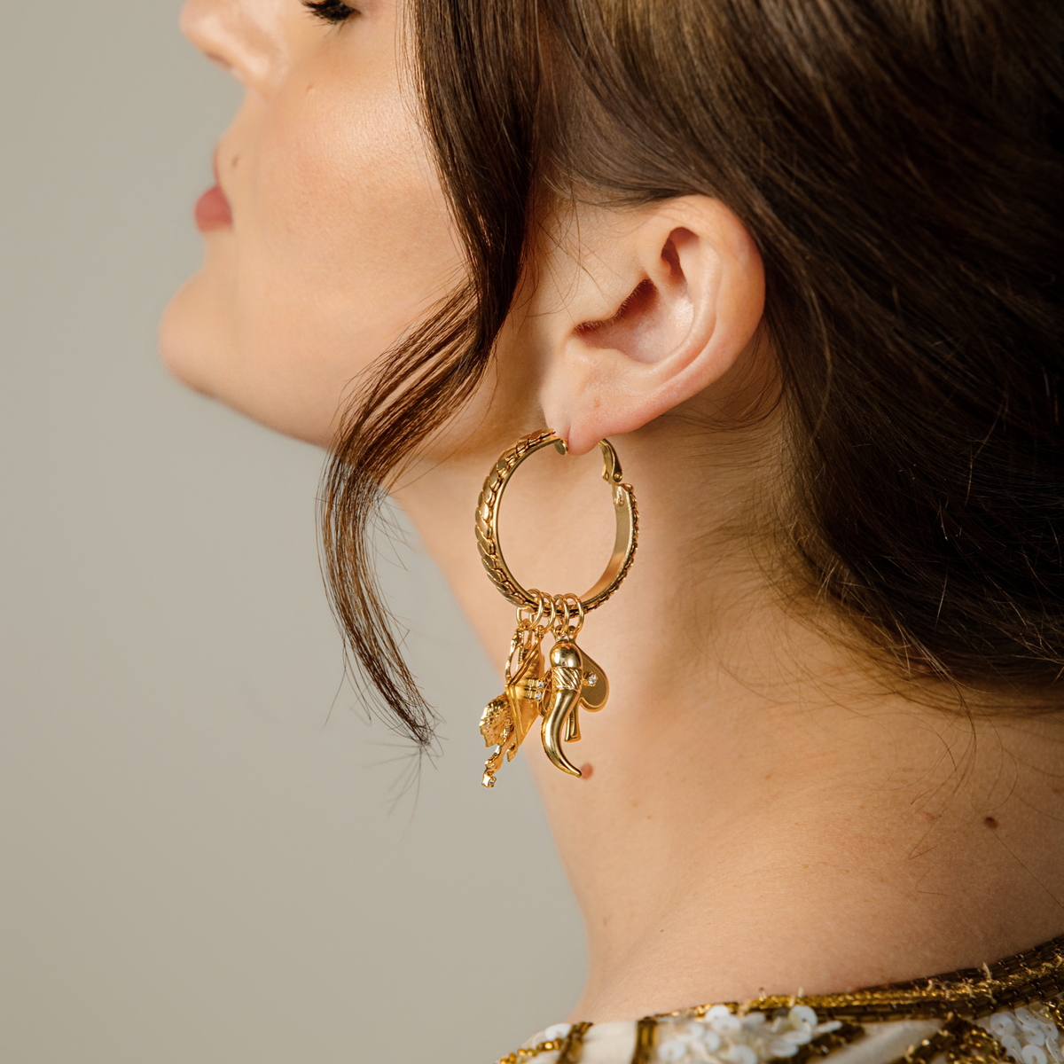 A side profile of a woman wearing gold-toned hinge hoop earrings with vintage chain details with four removable charms: lion, diamond, chili pepper, and spade with clear Swarovski crystal accents.