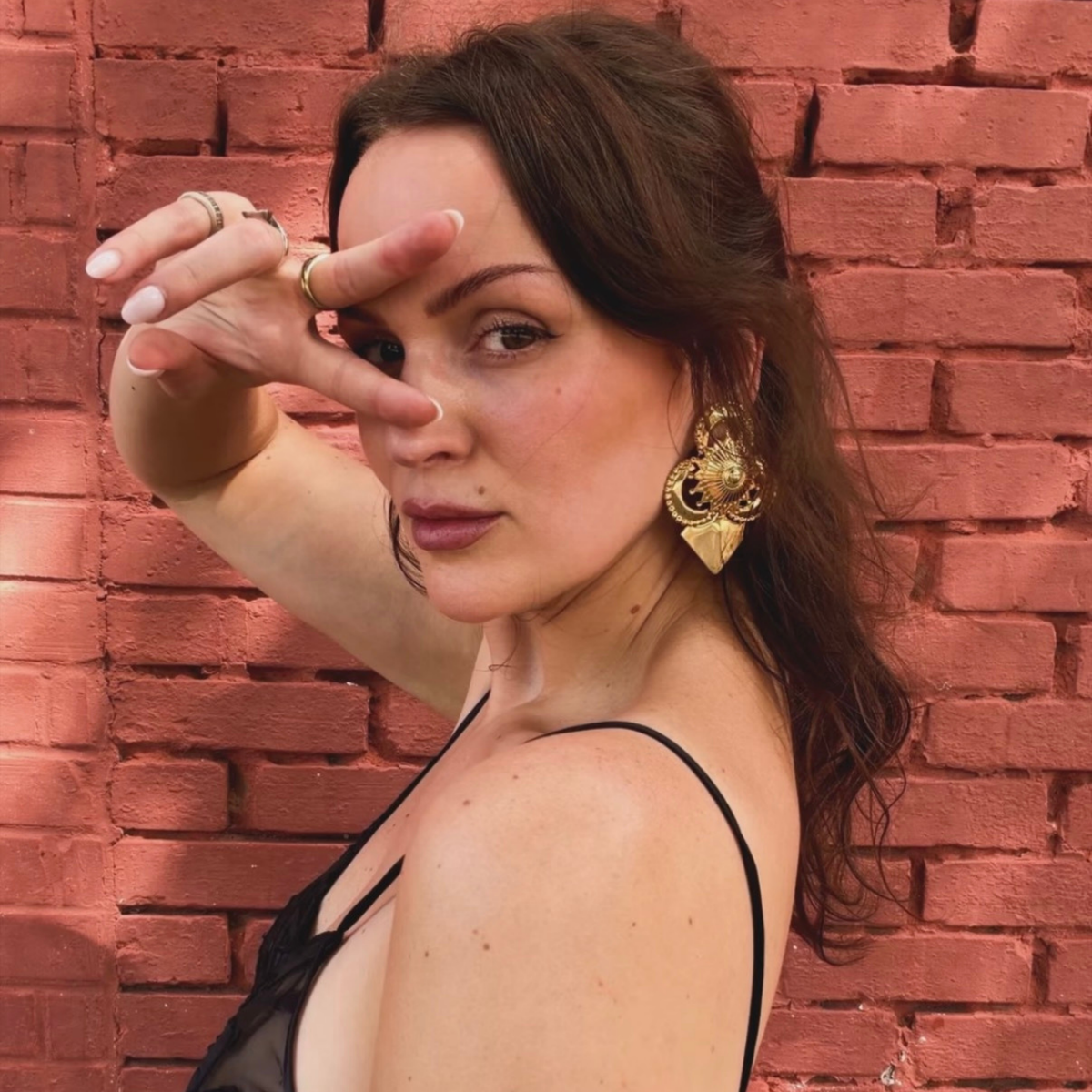 A woman wearing a pair of gold-toned geometric stud earrings with intricate detail, and a pointed triangular base against a red brick wall.