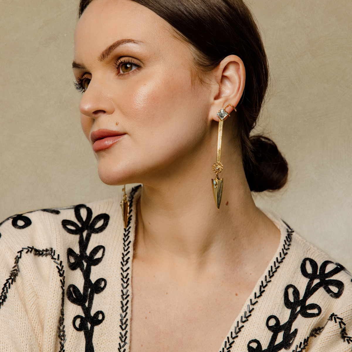 A close-up of a woman wearing a pair of gold-toned dangle snake chain earrings with attached faux cuff and clear Swarovski crystal accents, and pointed arrowhead charm at the bottom paired with a cream-colored embroidered top against a neutral beige background.