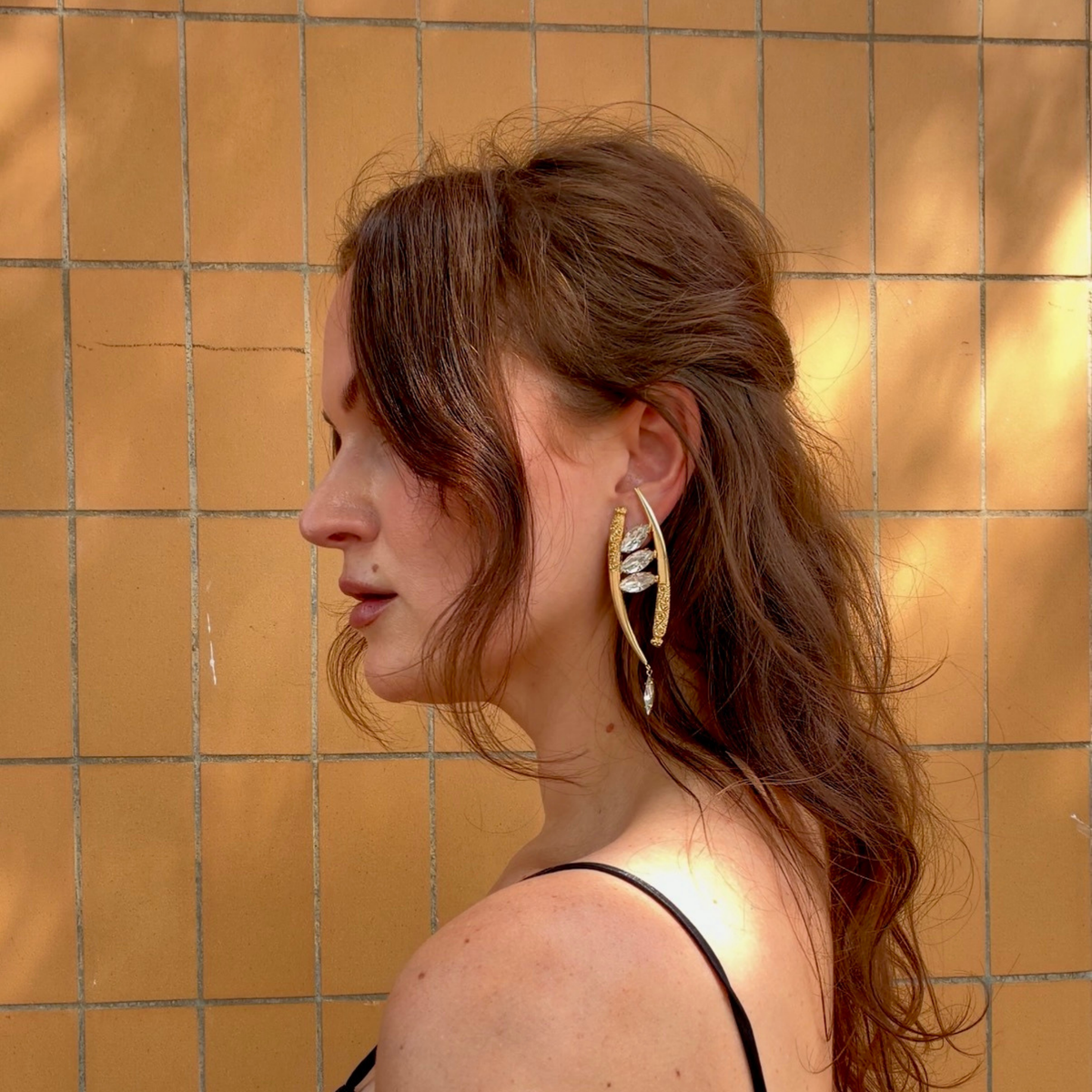 A woman in profile wearing a pair of asymmetric gold crescent-shaped drop earrings featuring marquise-cut clear, Swarovski crystals and ornate gold filigree details, set against an orange background.