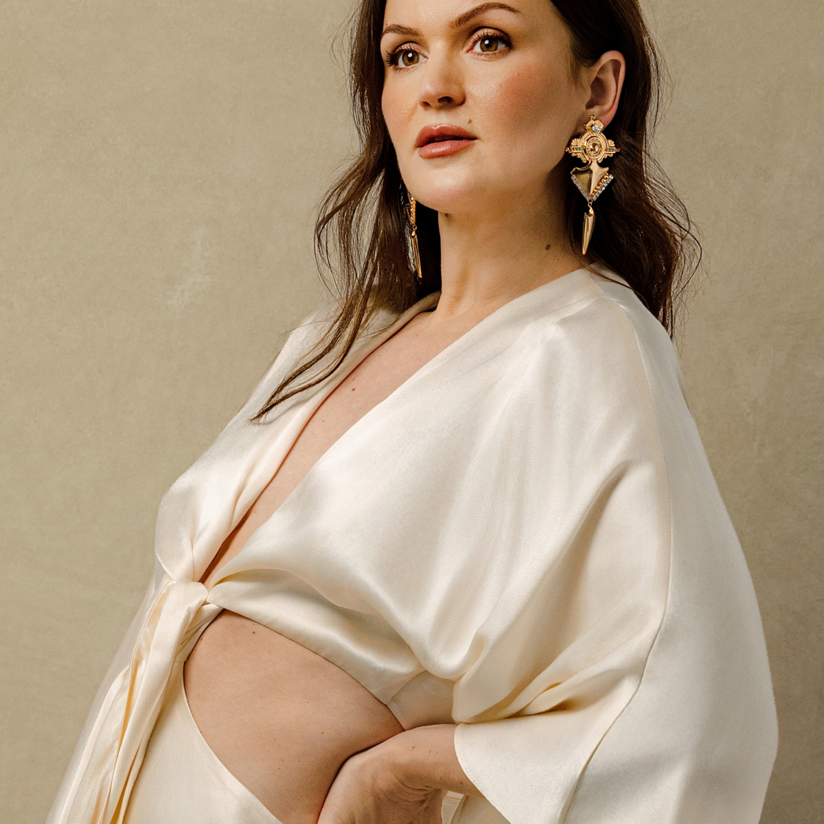 A model wearing a cream satin dress and large, ornate gold statement earrings and posing against a textured beige background.