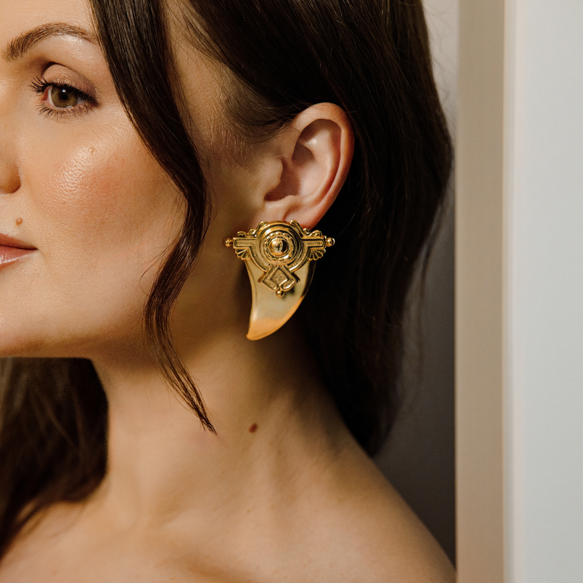 A close-up side profile of a woman wearing a large, ornate gold-toned shark tooth-shaped stud earrings with geometric detail.