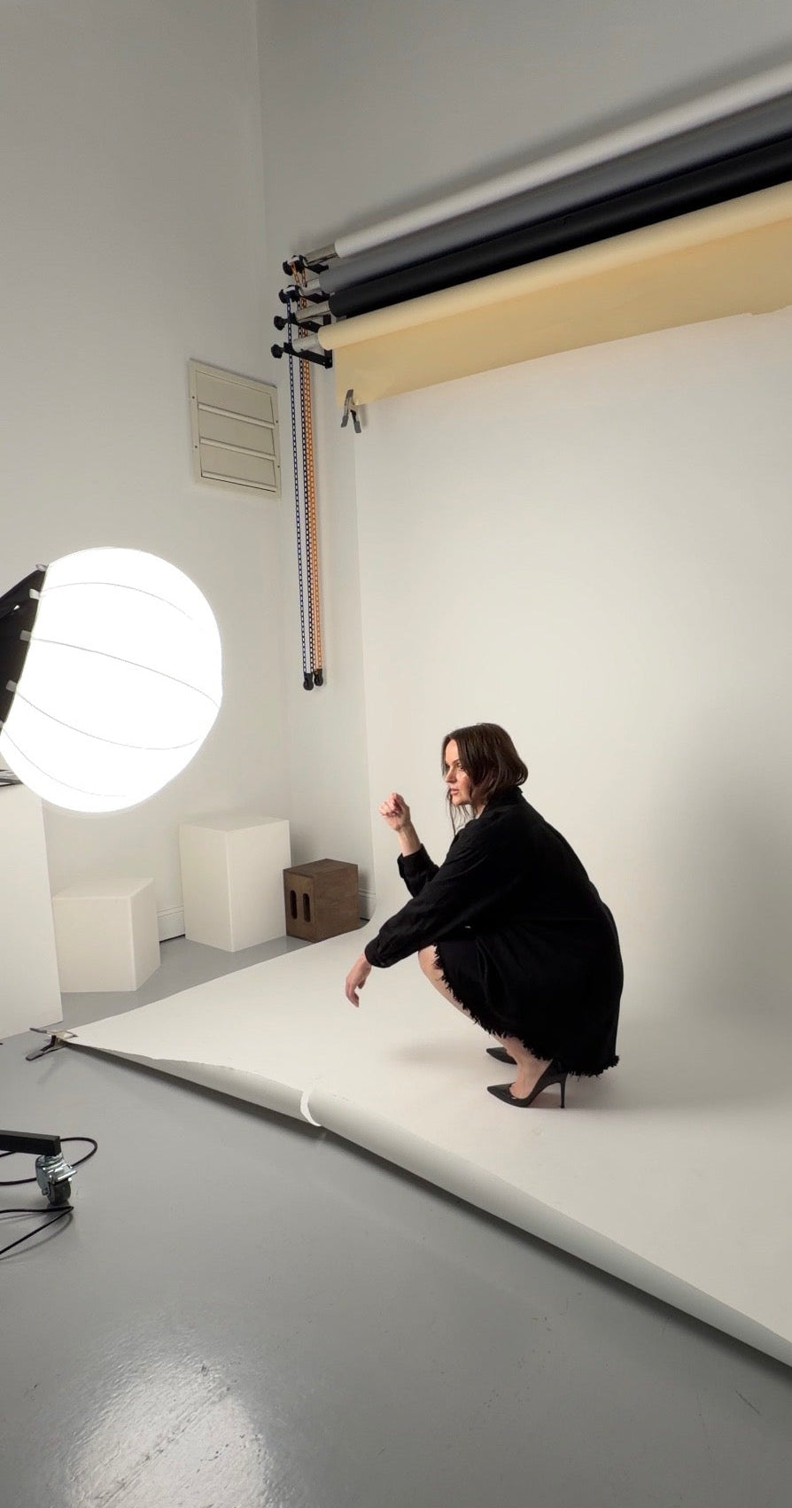 A woman in a black outfit crouches on a large white studio backdrop, with studio lighting equipment showing statement earrings.