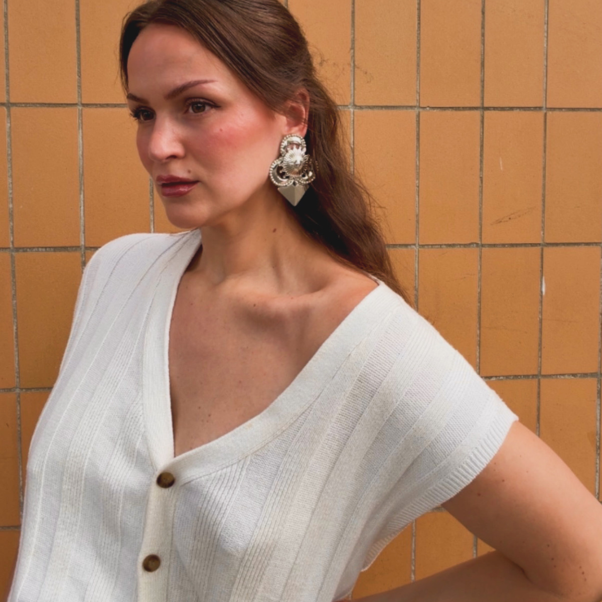 A woman wearing a pair of silver-toned geometric stud earrings with intricate detail, and a pointed triangular base against a terracotta tile wall.