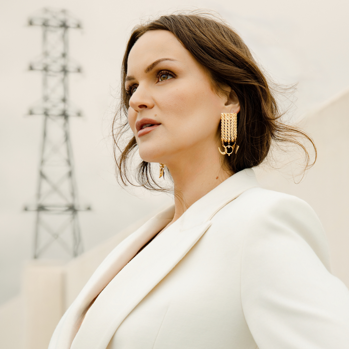A woman in a white blazer wearing a pair of chunky gold hoop earrings with a textured, layered chain-like design with horseshoe, hoop, and shield mini charms. She is wearing a white blazer, and the background is a soft, out-of-focus neutral indoor setting., photographed outdoors against a soft, light background with a faint power line visible.