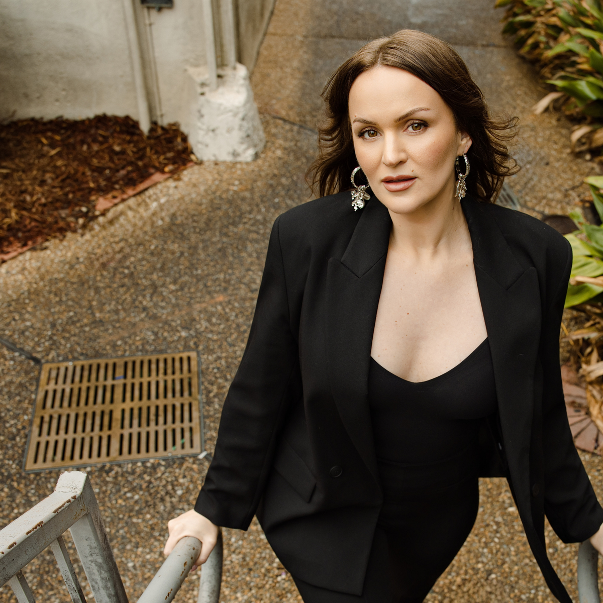A woman wearing silver-toned hinge hoop earrings with vintage chain details with four removable charms: lion, diamond, chili pepper, and spade with clear Swarovski crystal accents in an alley.
