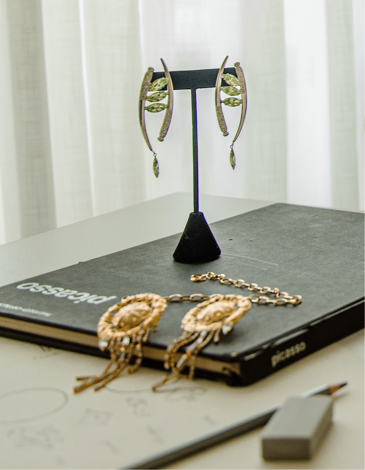 Gold-toned statement earrings with green leaf-like inlays and dangling teardrop charms, displayed on a black stand on top of a dark book, with matching gold-toned fringe jewelry and a pencil nearby on a light tabletop.