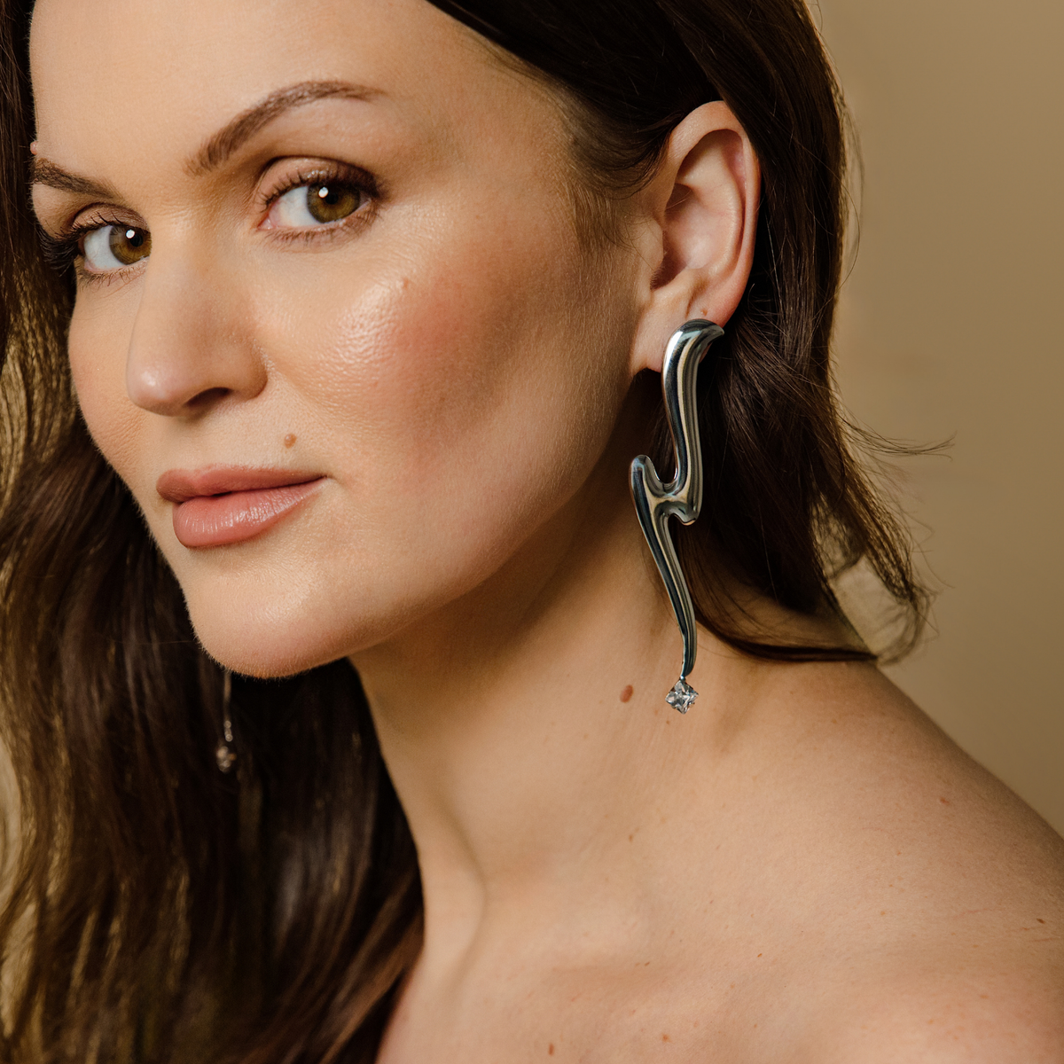 A close-up portrait of a woman wearing a dramatic, sculptural silver earring with a curved, lightning-bolt-like design and a small Swarovski crystal at the bottom.