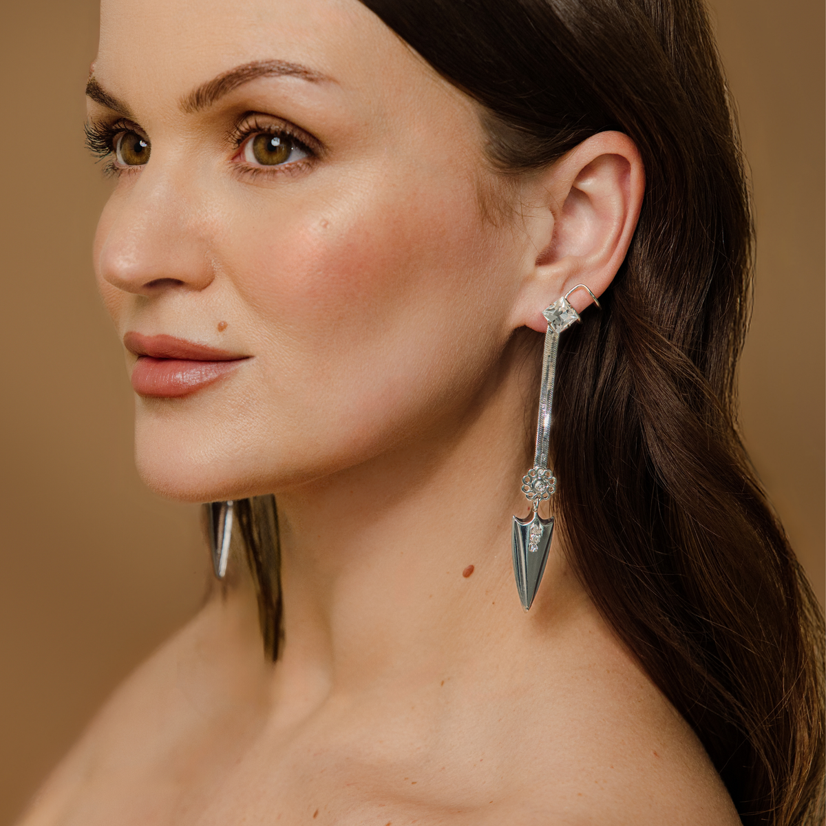 A close-up of a woman wearing a pair of silver-toned dangle snake chain earrings with attached faux cuff and clear Swarovski crystal accents, and pointed arrowhead charm at the bottom against a warm beige background.