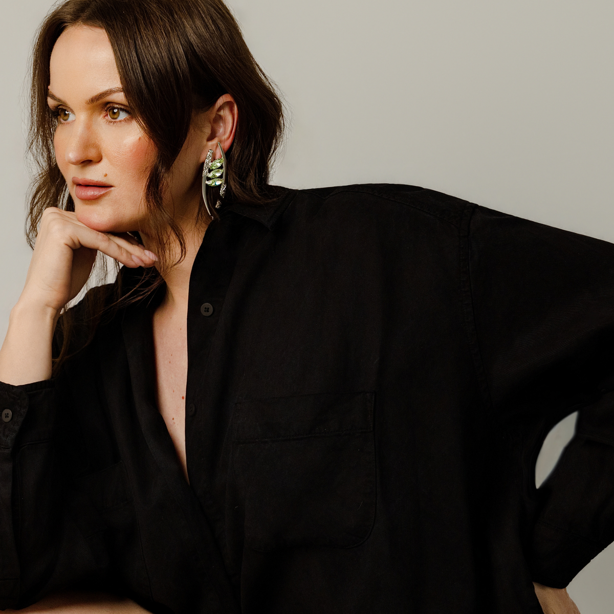 A woman wearing a black button-up shirt and a pair of silver-toned crescent-shaped drop earrings featuring marquise-cut mint green Swarovski crystals and ornate filigree details, posing against a neutral light gray background.