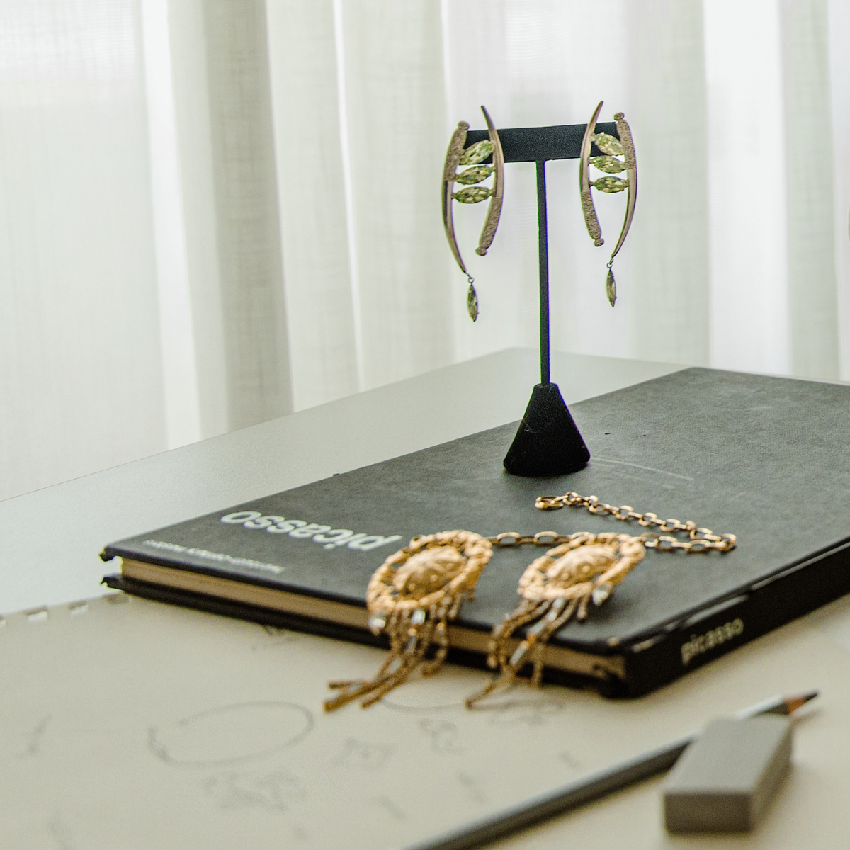 Gold-toned statement earrings with a pair of silver-toned crescent-shaped drop earrings featuring marquise-cut mint green Swarovski crystals and ornate filigree details displayed on a black stand on top of a dark book, with sketching supplies.