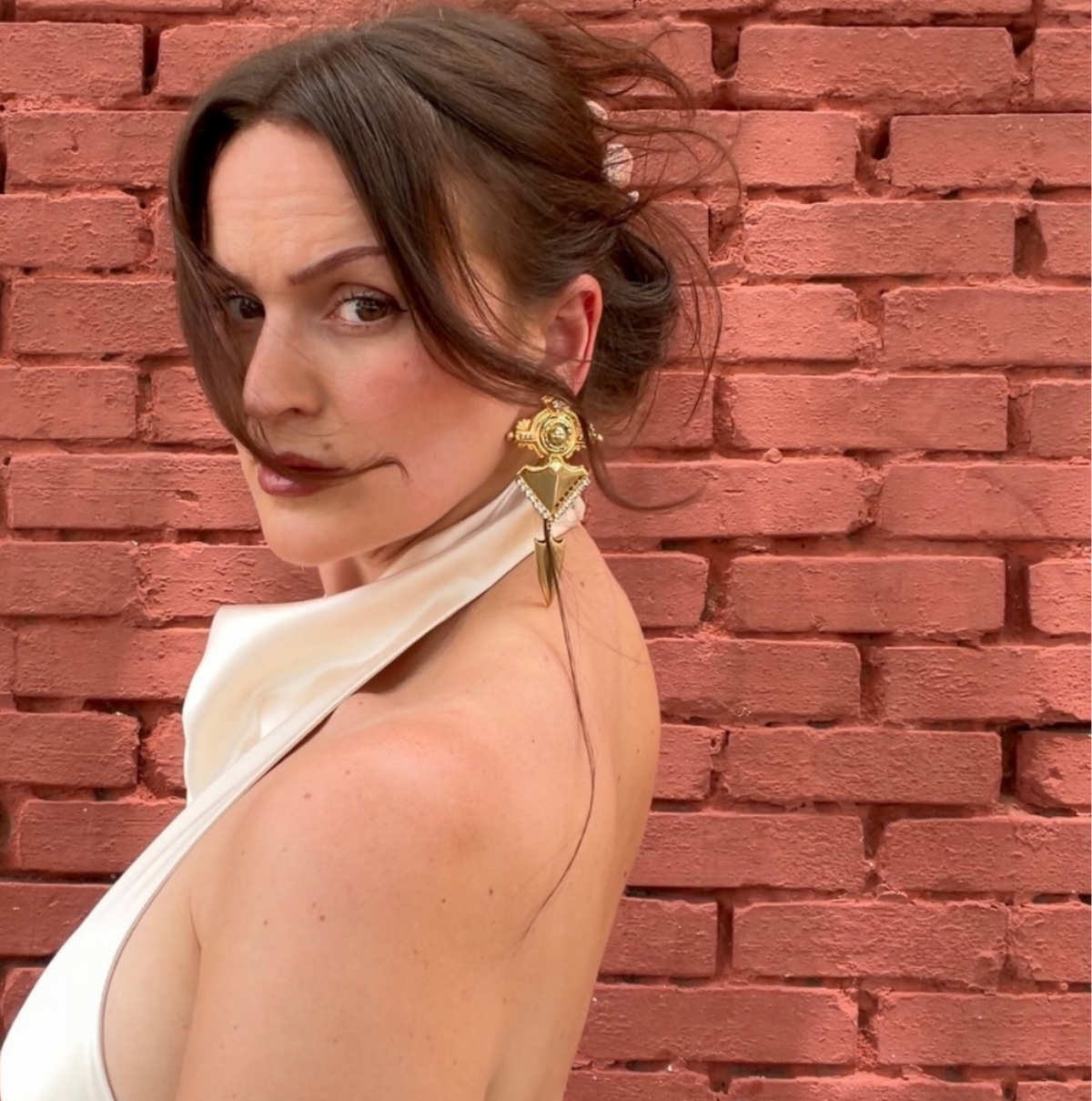 A woman with brown hair in a low bun wearing a white halter top and a large gold statement earring, posing in front of a red brick wall.