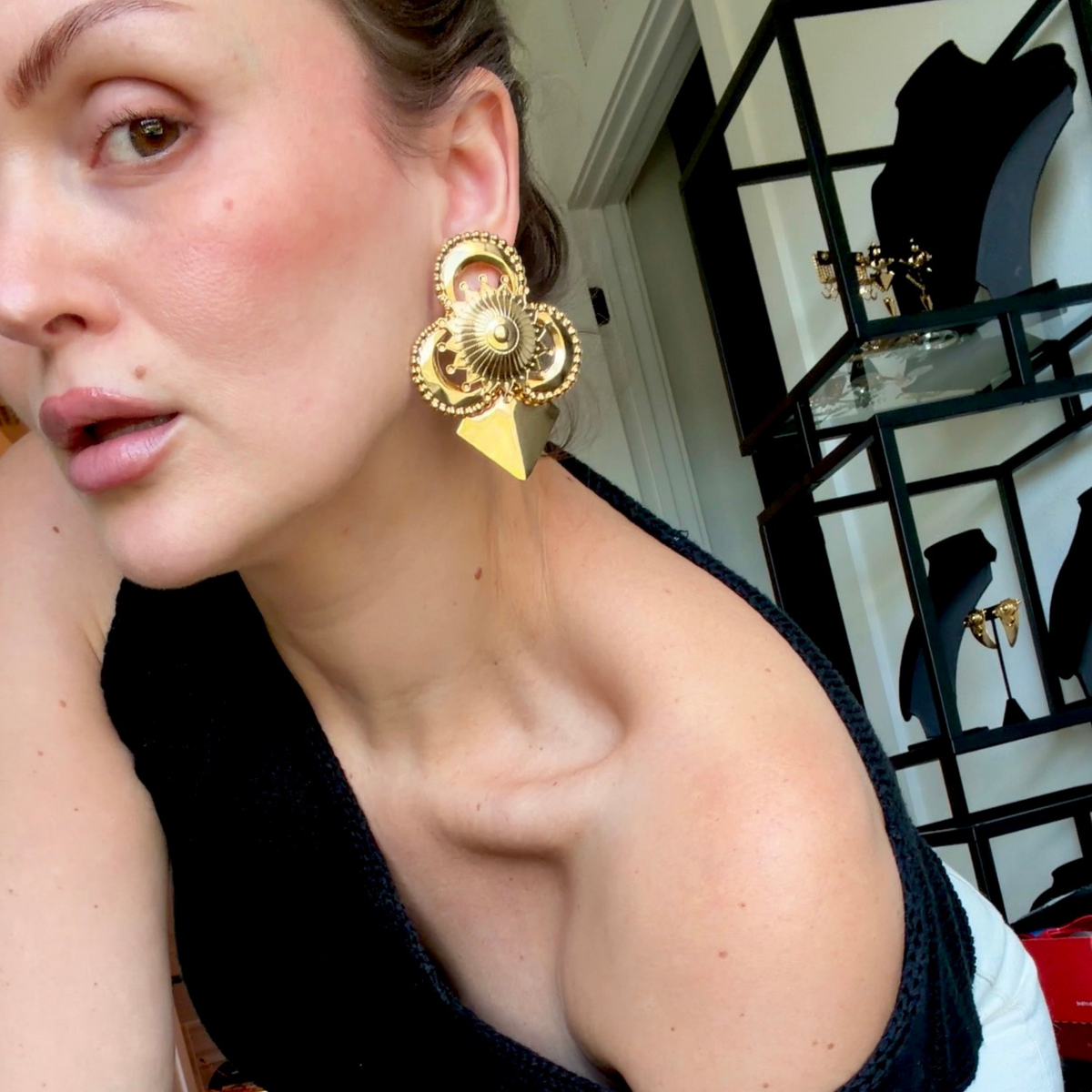 A woman in a black top wearing a pair of gold-toned geometric stud earrings with intricate detail, and a pointed triangular base.