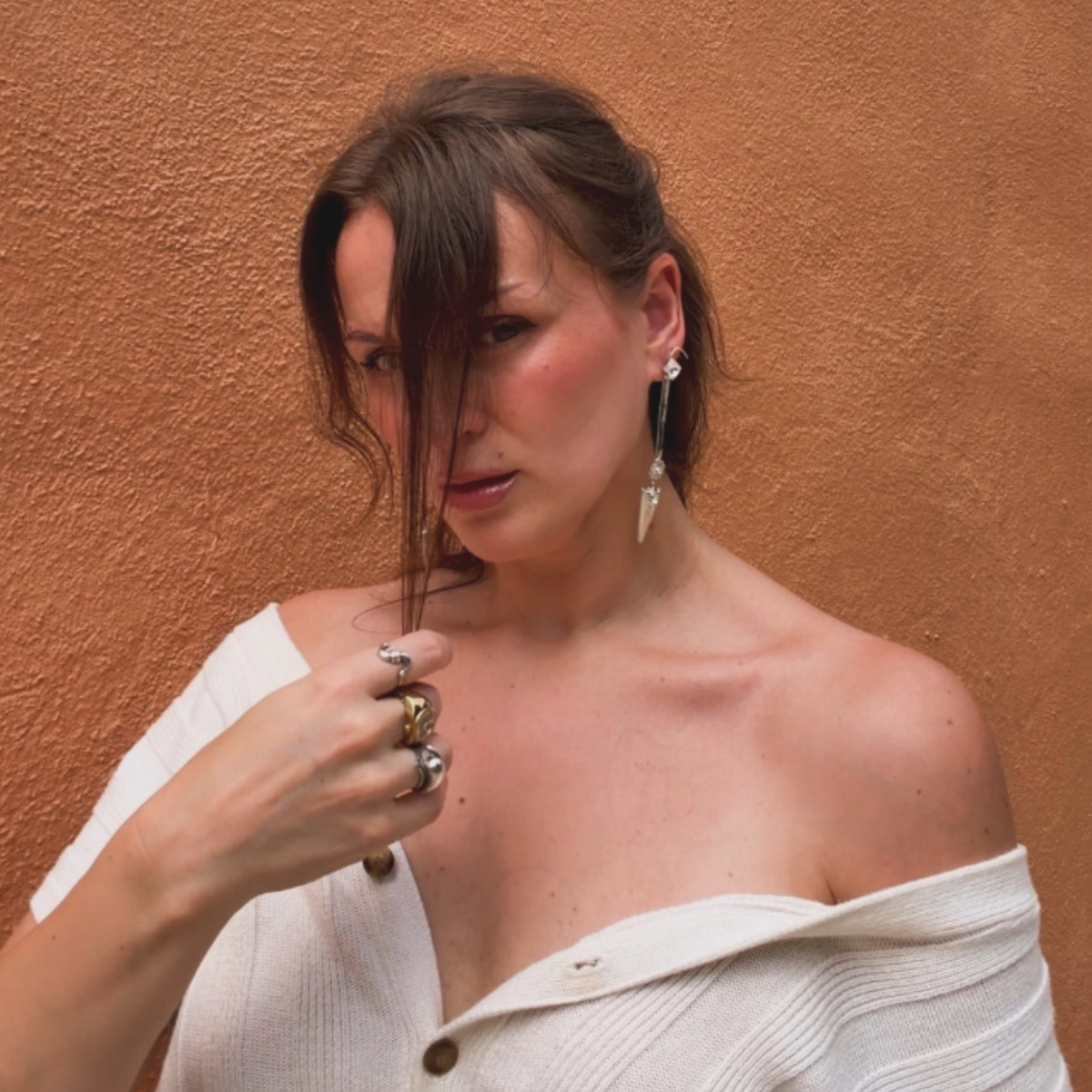 A woman wearing a white off-the-shoulder top and pair of silver-toned dangle snake chain earrings with attached faux cuff and clear Swarovski crystal accents, and pointed arrowhead charm at the bottom against a textured terracotta wall.