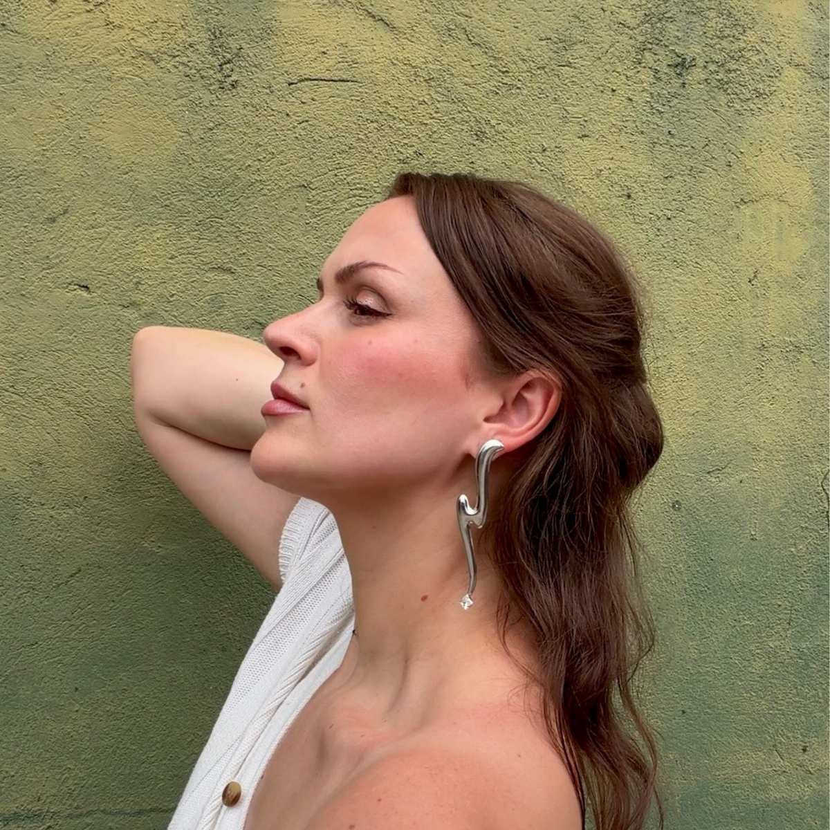 A woman wearing a long, sculptural silver earring with a clear Swarovski crystal at the bottom posing against a textured olive green wall.