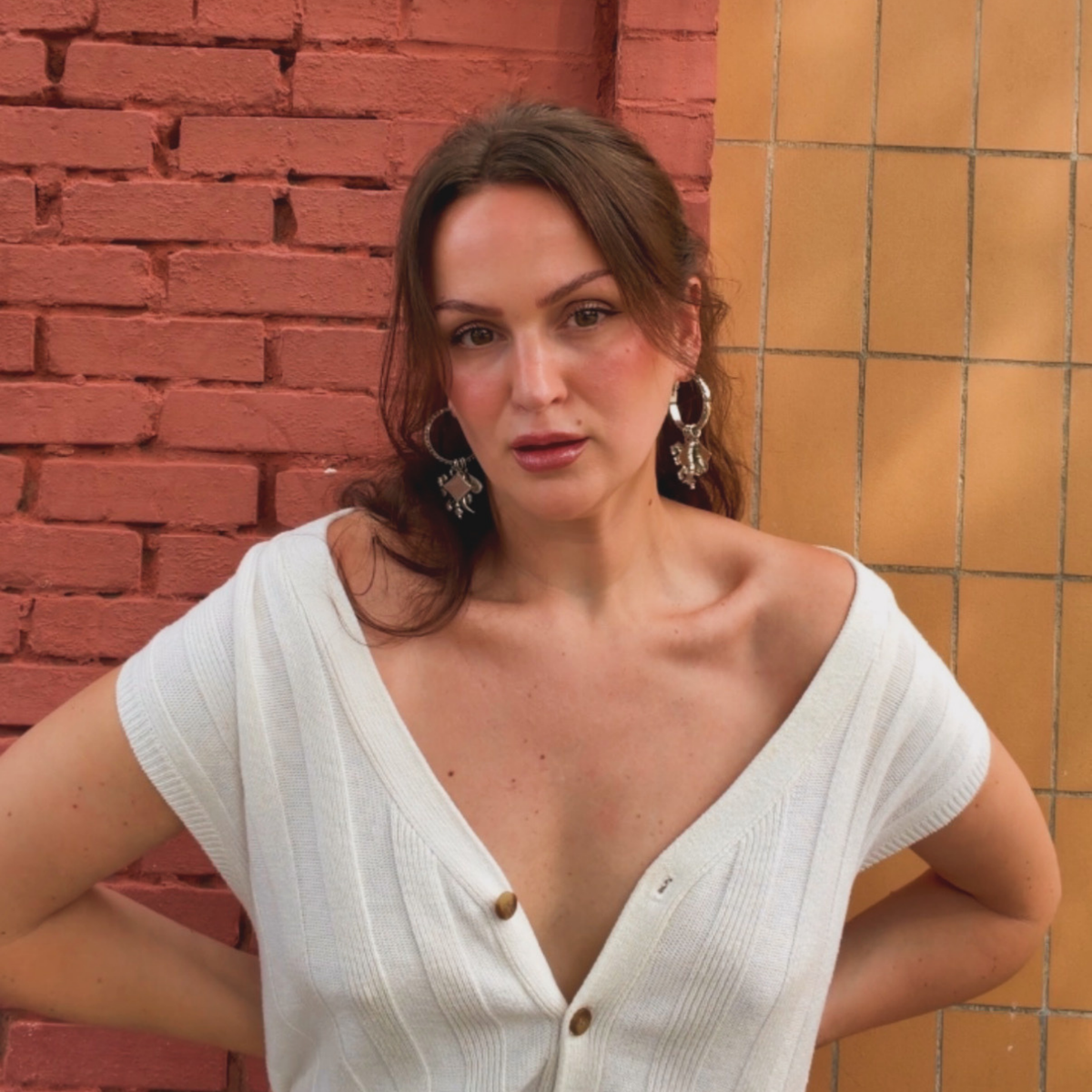 A woman wearing silver-toned hinge hoop earrings with vintage chain details with four removable charms: lion, diamond, chili pepper, and spade with clear Swarovski crystal accents against a brick wall.