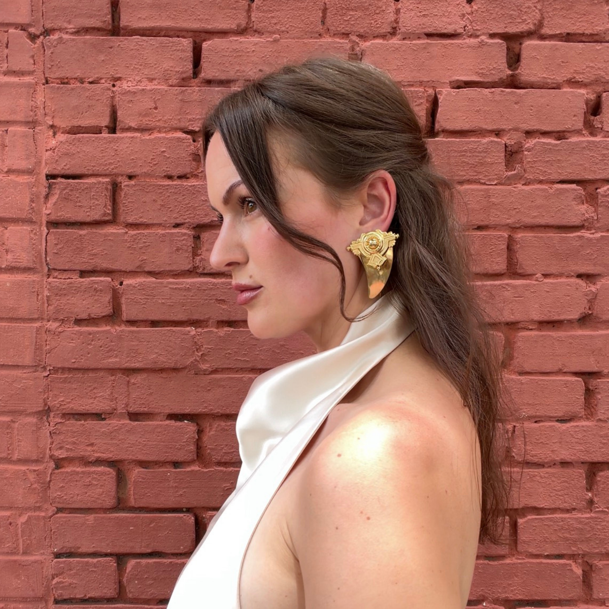 A woman in profile wearing large, gold-toned shark tooth-shaped stud earrings with geometric detail against a red brick wall background.
